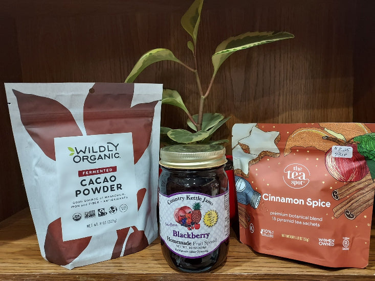 A small fall-themed display featuring a bag of Wildly Organic fermented cacao powder, a jar of Country Kettle blackberry fruit spread, and a box of The Tea Spot cinnamon spice tea arranged on a wooden shelf with a green potted plant behind them.