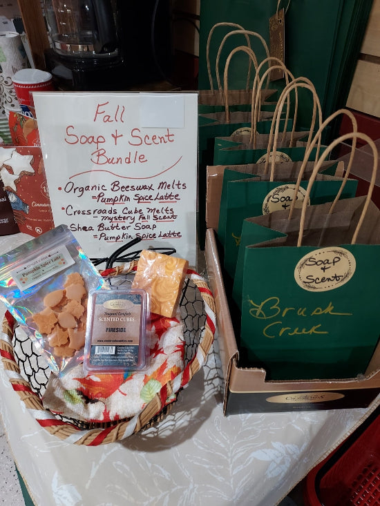 A fall soap and scent bundle display featuring a basket with a scented candle, beeswax melts, and a small packaged item, alongside multiple green gift bags labeled 'Brush Creek' and 'Soap + Scent' on a table.