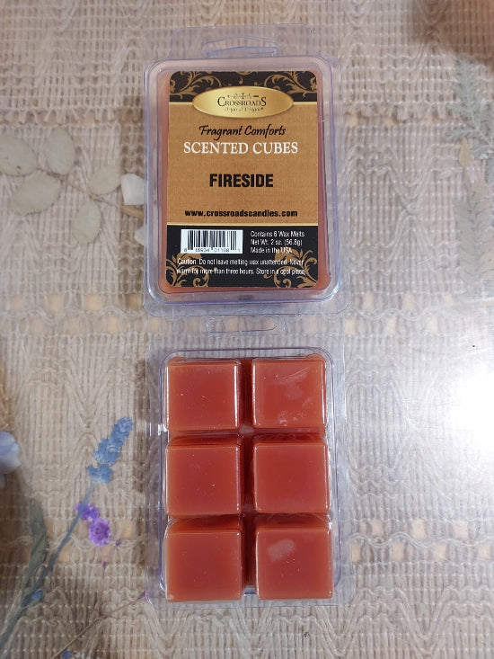 A package of Crossroad Scents 'Fireside' scented wax melts shown above an open clear plastic clamshell containing six reddish-brown wax cubes, placed on a textured beige fabric surface.