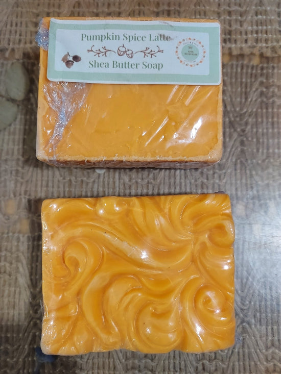 Two orange-yellow soap bars on a textured brown surface. The top bar is wrapped in clear plastic with a label reading 'Pumpkin Spice Latte Shea Butter Soap.