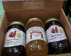 Three glass jars of fruit spread arranged inside a brown cardboard gift box. The jars are labeled Country Kettle Jams: Cherry Berry, Grandma's Jam House Pineapple Jam, and Blackberry.