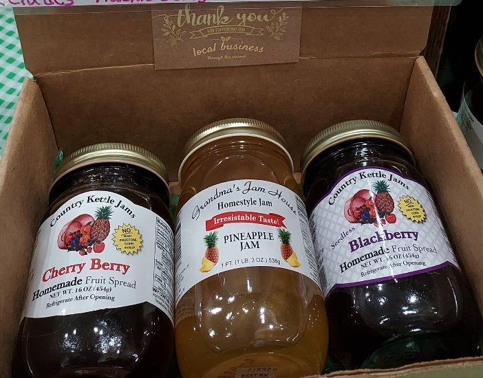 Three glass jars of fruit spread arranged inside a brown cardboard gift box. The jars are labeled Country Kettle Jams: Cherry Berry, Grandma's Jam House Pineapple Jam, and Blackberry.