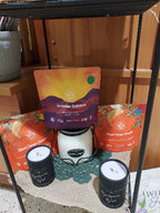 A collection of tea products from the brand Tea Time Tales, including two black metal candle jars with white lids, a white candle in a glass container, and two colorful tea bags (Orange Blossom and Summer Solstice) on a burlap runner over a wooden shelf.