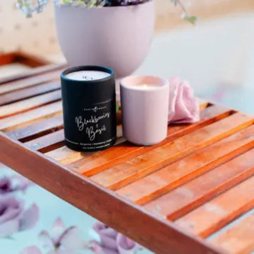 Blackberry Basil soy candle on a wooden slatted tray, featuring a matte black jar sleeve labeled 'Blackberry Basil' and a matte pale pink ceramic candle, styled with a pink rose and a light purple planter in the background.
