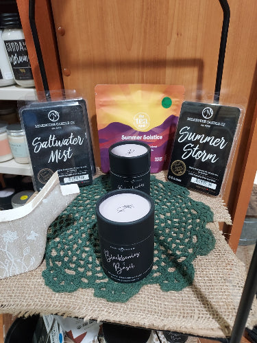 A set of three Bath Body Works scented products arranged on a wooden table: a Summer Storm shower foam stick, a Saltwater Mist bar, and a Blackwater Bath Bomb stick, alongside a colorful insert card and a green crocheted doily.