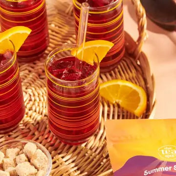 A close-up of a glass of red iced beverage with a clear straw and orange slice garnish, set on a woven tray with other similar glasses, sugar cubes, and a yellow packet labeled Summer S.