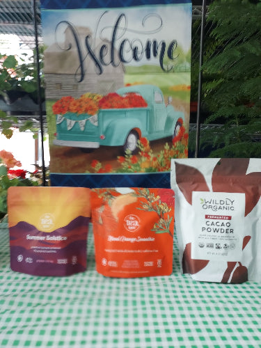 A collection of packaged food products arranged on a green-and-white checkered surface, including a decorative sign with a teal truck and flowers, a bag of Valdity Cacao Powder, an orange package, and a purple package.