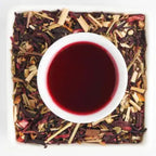 A top-down view of a white cup filled with dark red tea, surrounded by loose-leaf herbal tea ingredients including dried flowers, stems, and wood pieces on a white square plate.