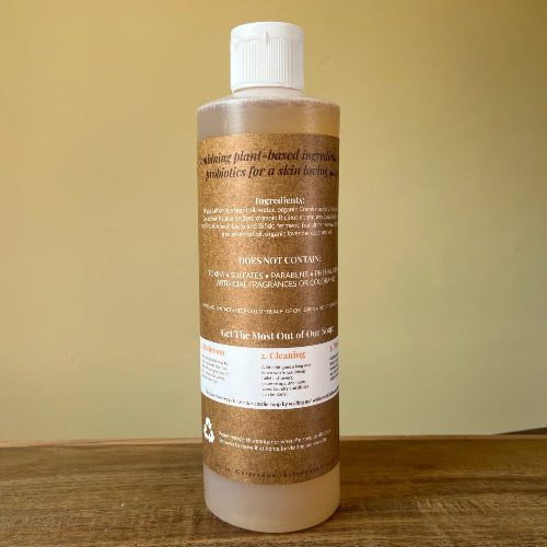 A bottle of Plant Based Invigorators Probiotics for a skin loving body wash, shown from the back on a wooden surface against a beige background.