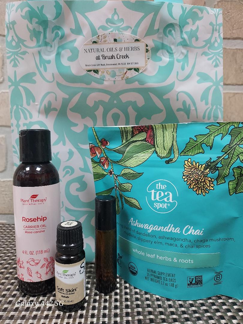 A tea and essential oil gift set featuring a large patterned tote bag, a bag of Ashwagandha Chai herbal tea, a bottle of rosehip cosmetic oil, and two small amber glass bottles, arranged on a checkered surface in front of a tiled wall.