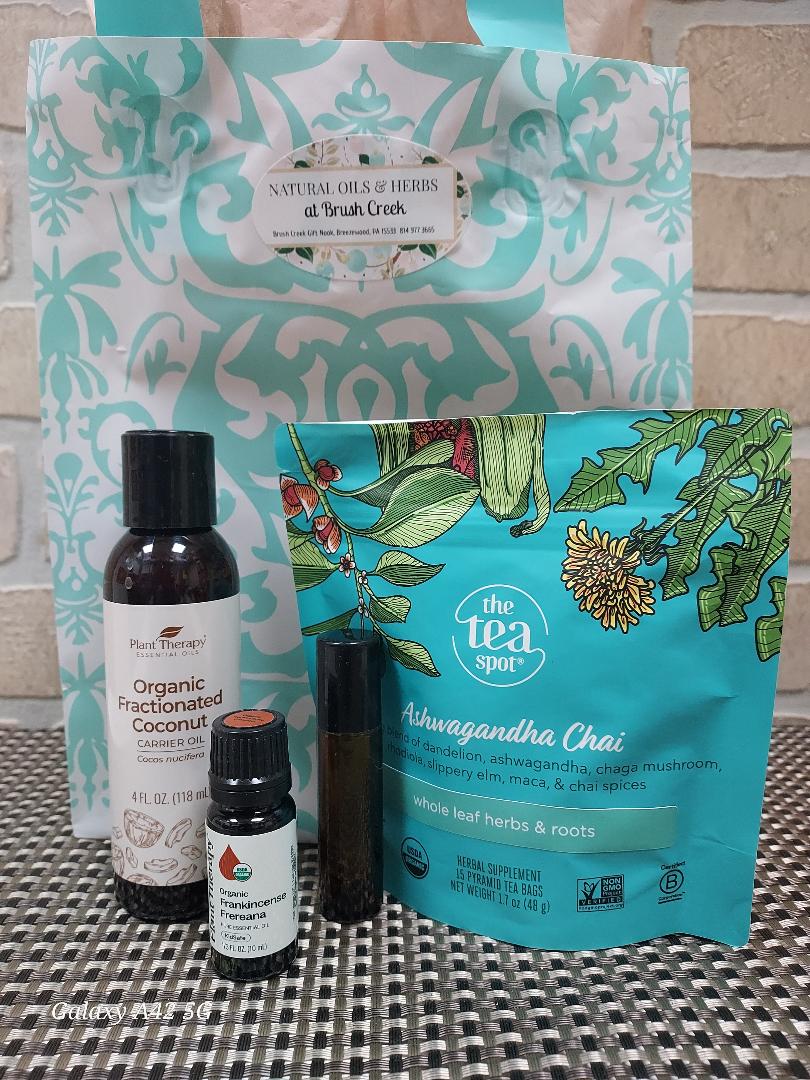 A tea and essential oil gift set featuring a large teal and white patterned bag from Natural Oils of Herbs by Brush Creek, a teal stand-up pouch of The Tea Spot Ashwagandha Chai, and two dark amber bottles of essential oils on a black-and-white checkered surface.