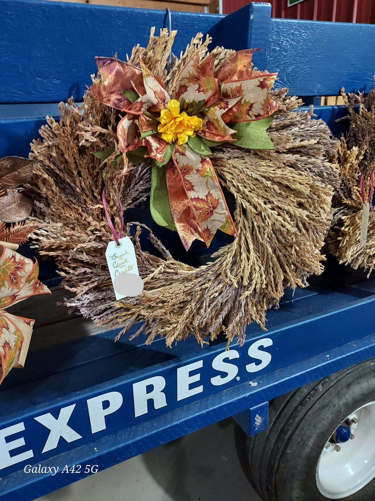 A rustic autumn wreath made of dried brown corn plumes, decorated with a large patterned bow in fall colors and a small yellow flower, displayed on a blue wagon.