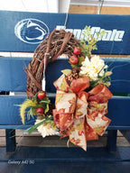 An autumn grapevine wreath with a large orange and beige patterned bow, white flowers, green foliage, and red berries, displayed on a blue Penn State bench.