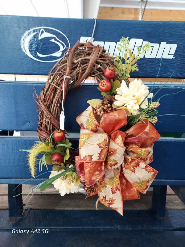 An autumn grapevine wreath with a large orange and beige patterned bow, white flowers, green foliage, and red berries, displayed on a blue Penn State bench.