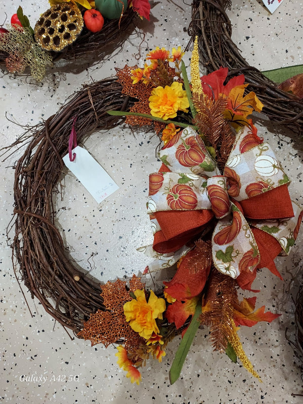 An autumn grapevine wreath decorated with a large fabric bow featuring a pumpkin pattern, orange faux leaves, yellow flowers, and metallic gold accents, displayed on a speckled light gray countertop.