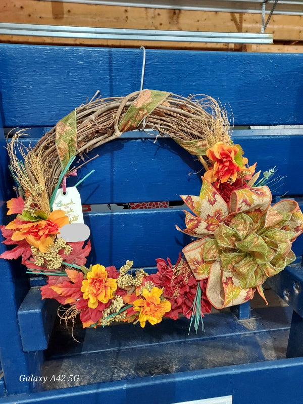 An autumn-themed grapevine wreath with artificial orange, yellow, and red leaves, a large patterned bow, and dried grass accents, displayed on a blue wooden crate.
