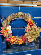 An autumn-themed grapevine wreath with artificial orange, yellow, and red leaves, a large patterned bow, and dried grass accents, displayed on a blue wooden crate.