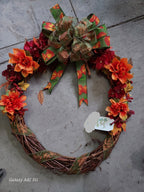 An autumn-themed grapevine wreath with orange and red artificial flowers, green and orange patterned ribbon accents, and a large matching bow on top, photographed on a gray stone floor.