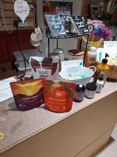 A collection of assorted products, including several pouches of coffee or tea, a white mug, and small bottles, arranged on a patterned surface next to a large brown cardboard box in a cluttered indoor setting.