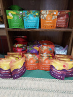 Multiple pouches of tea and herbal blends arranged on a shelf and tabletop. The front row shows several orange, yellow, and purple pouches labeled 'Sunflower Blossom' and 'Hand Orange Herbal', while the shelves behind display other colorful packages including green, blue, and orange variants.